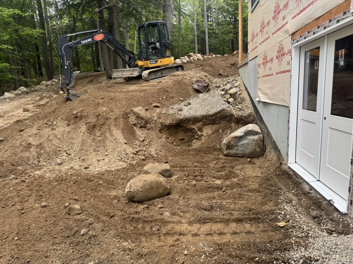 Mini excavator grading beside a new home foundation with boulder wall